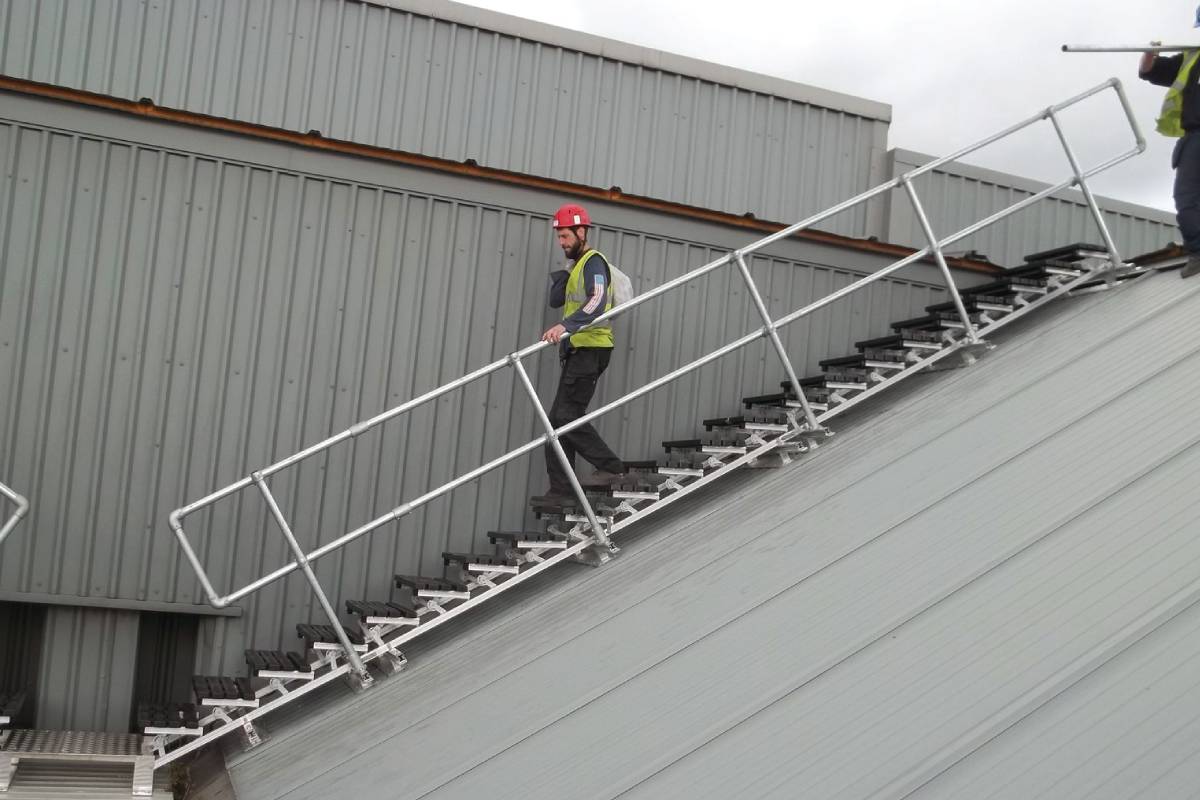 Operario con casco y chaleco de seguridad descendiendo por una escalera técnica con barandilla en una cubierta inclinada